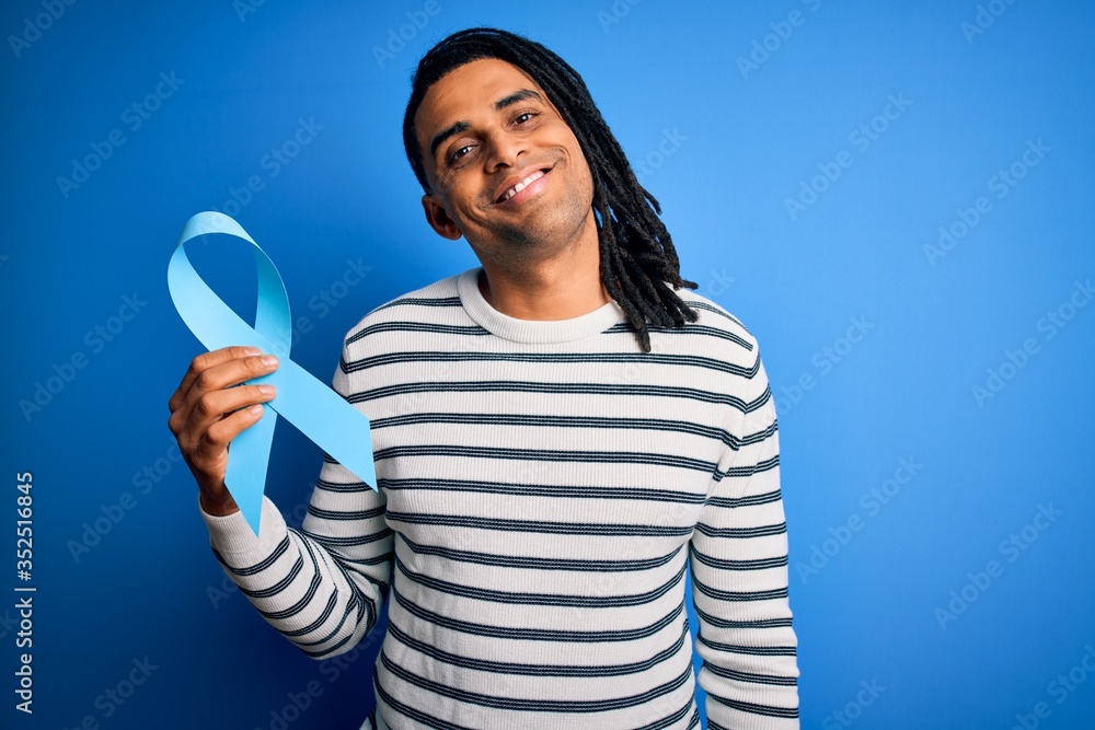 Young african american afro man with dreadlocks holding blue cancer ribbon with a happy face standing and smiling with a confident smile showing teeth Young african american afro man with dreadlocks holding blue cancer ribbon with a happy face standing and smiling with a confident smile showing teeth
