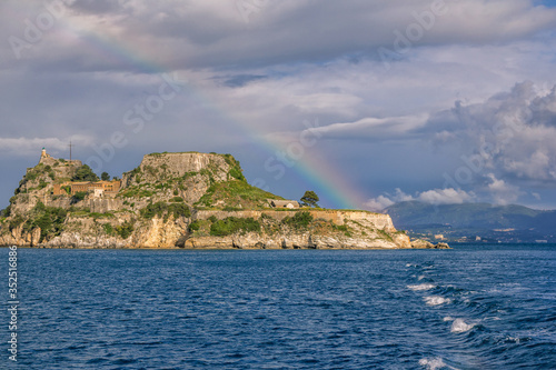 View of ancient venetian fortress on the hill at Corfu Island, Greece. Old stone walls, covered with green grass, sea bay, blue sky and rainbow