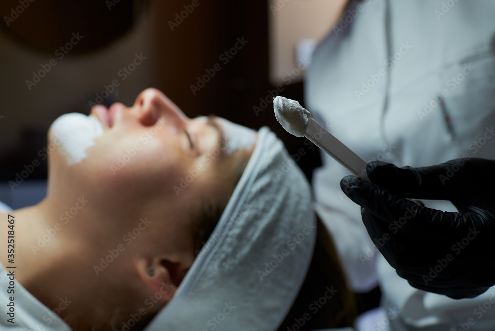 A close-up photo of peeling gommage on a stick near a young woman's face on a beauty bed. A cosmetologist wearing black disposable medical gloves during a procedure in a beauty salon for skin cleaning