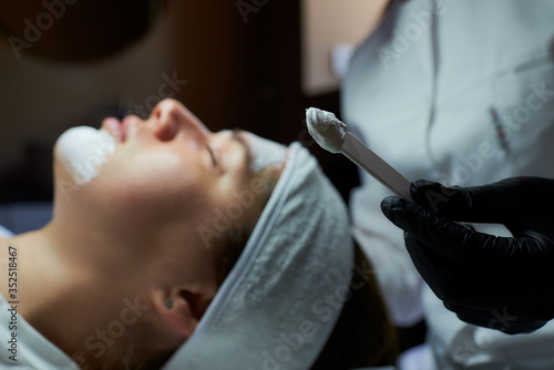 Obraz na płótnie A close-up photo of peeling gommage on a stick near a young woman's face on a beauty bed. A cosmetologist wearing black disposable medical gloves during a procedure in a beauty salon for skin cleaning w salonie