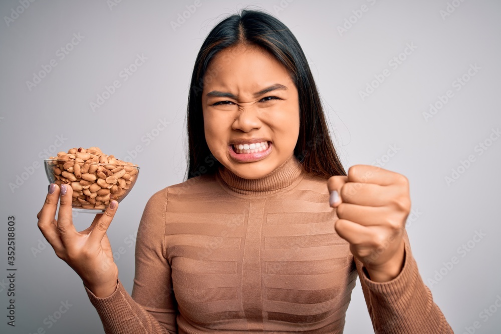 Young asian girl holding bowl of healthy peanuts eating as snack for diet over white background annoyed and frustrated shouting with anger, crazy and yelling with raised hand, anger concept Young asian girl holding bowl of healthy peanuts eating as snack for diet over white background annoyed and frustrated shouting with anger, crazy and yelling with raised hand, anger concept