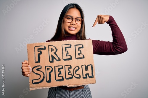 Asian girl asking for rights holding banner with free speech message over white background with surprise face pointing finger to himself
