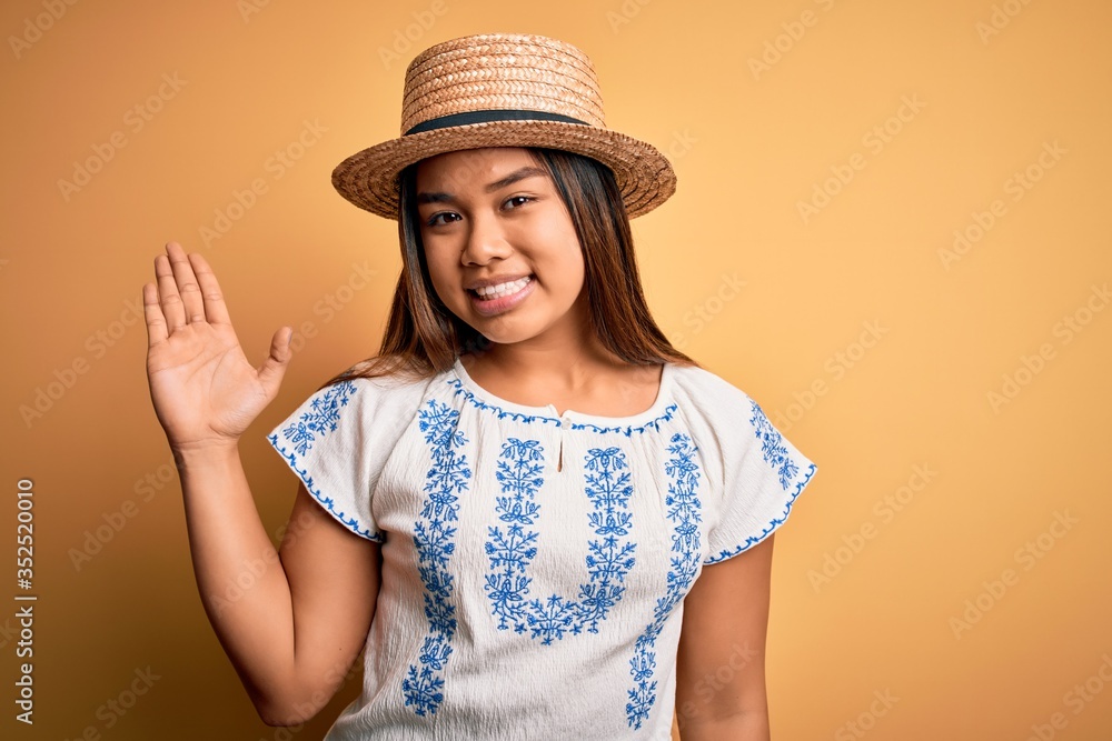 Young beautiful asian girl wearing casual t-shirt and hat standing over yellow background Waiving saying hello happy and smiling, friendly welcome gesture Young beautiful asian girl wearing casual t-shirt and hat standing over yellow background Waiving saying hello happy and smiling, friendly welcome gesture