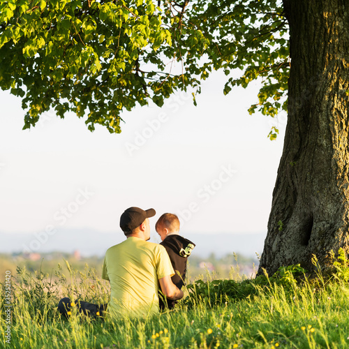Dad with son in the spring meadow sitting under the tree in tall grass. Travel with child concept