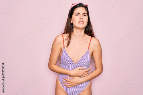 Young beautiful fashion girl wearing swimwear swimsuit and sunglasses over pink background with hand on stomach because indigestion  painful illness feeling unwell. Ache concept.