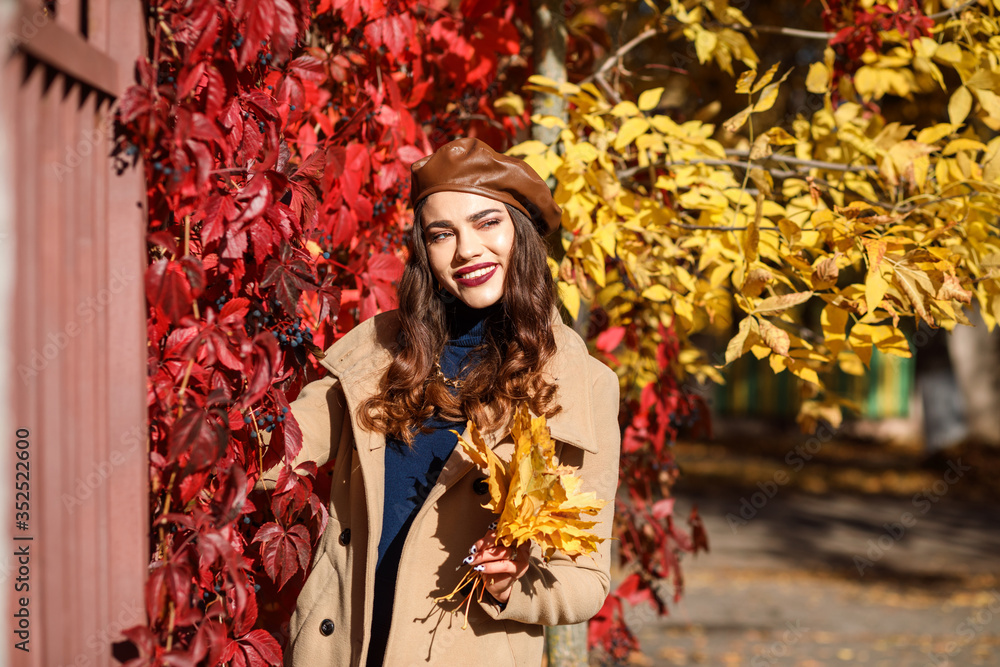 Cheerful smiling woman is spending time outdoors in autumn. Caucasian woman is wearing brown leather cap and beige coat. Red and yellow leaves in the background. Stylish girl. 