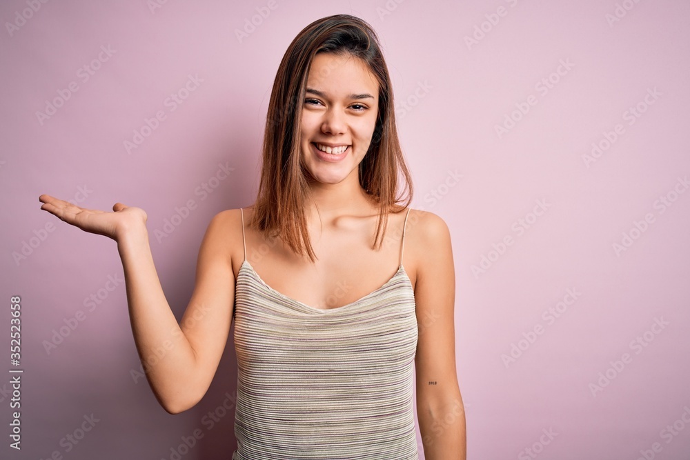 Young beautiful brunette girl wearing casual striped t-shirt over isolated pink background smiling cheerful presenting and pointing with palm of hand looking at the camera. Young beautiful brunette girl wearing casual striped t-shirt over isolated pink background smiling cheerful presenting and pointing with palm of hand looking at the camera.