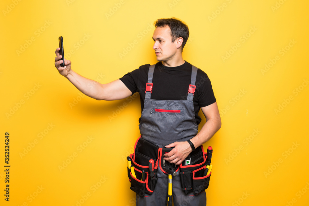 Young man worker with tools belt holding phone isolated on yellow background Young man worker with tools belt holding phone isolated on yellow background