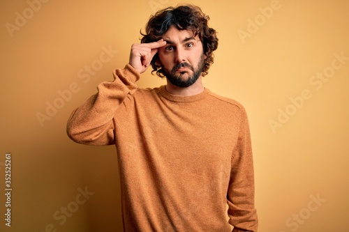 Young handsome man with beard wearing casual sweater standing over yellow background worried and stressed about a problem with hand on forehead, nervous and anxious for crisis