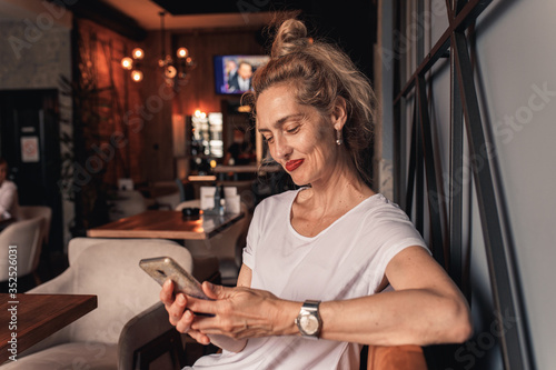 Smiling senior woman sitting in cafe drinking coffee and using smartphone.