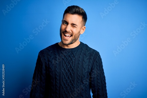 Photography Young handsome man wearing casual sweater standing over isolated blue background winking looking at the camera with sexy expression, cheerful and happy face