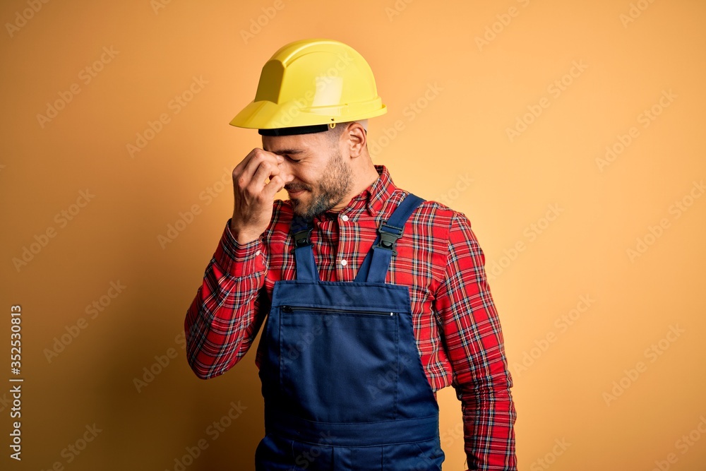 Young builder man wearing construction uniform and safety helmet over yellow isolated background tired rubbing nose and eyes feeling fatigue and headache. Stress and frustration concept. Young builder man wearing construction uniform and safety helmet over yellow isolated background tired rubbing nose and eyes feeling fatigue and headache. Stress and frustration concept.