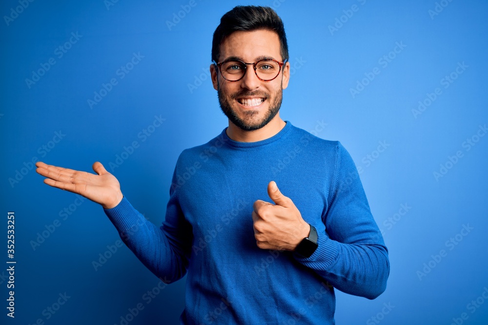 Young handsome man with beard wearing casual sweater and glasses over blue background Showing palm hand and doing ok gesture with thumbs up, smiling happy and cheerful Young handsome man with beard wearing casual sweater and glasses over blue background Showing palm hand and doing ok gesture with thumbs up, smiling happy and cheerful