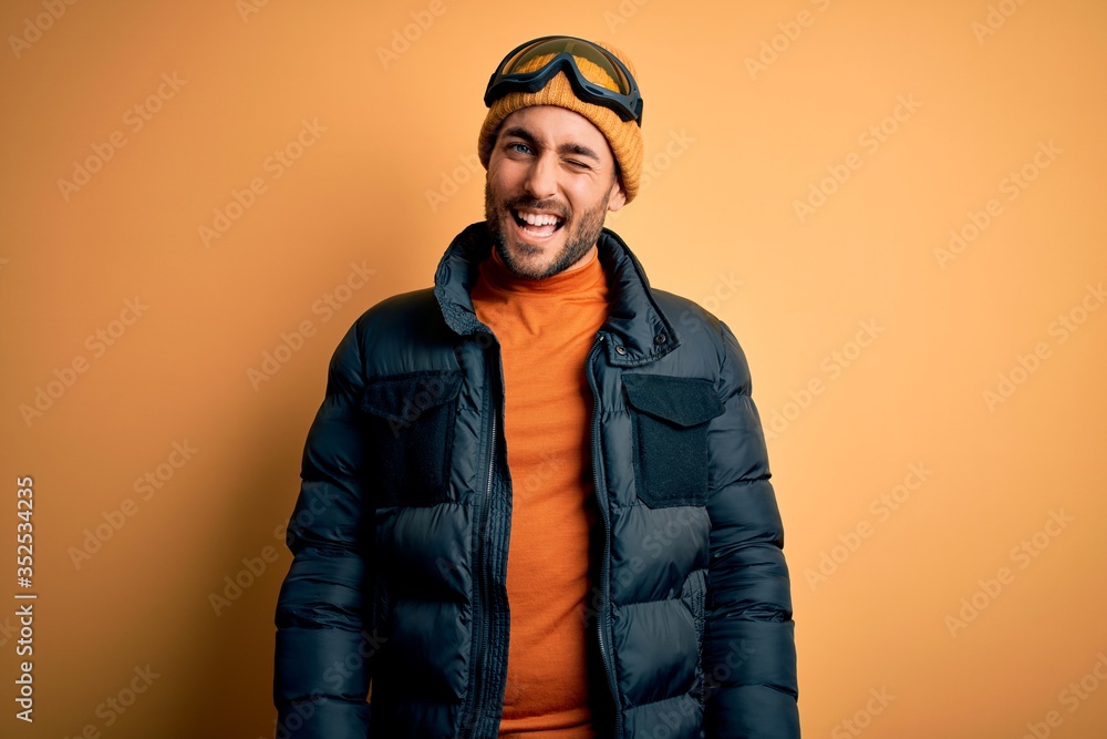 Young handsome skier man with beard wearing snow sportswear and ski goggles winking looking at the camera with sexy expression, cheerful and happy face. Young handsome skier man with beard wearing snow sportswear and ski goggles winking looking at the camera with sexy expression, cheerful and happy face.