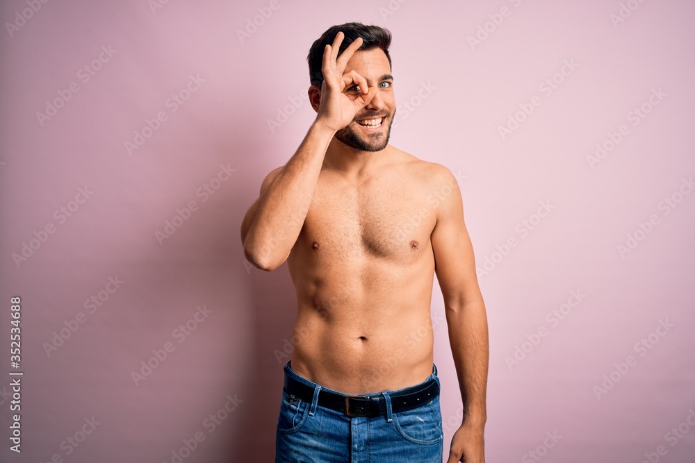 Young handsome strong man with beard shirtless standing over isolated pink background doing ok gesture with hand smiling, eye looking through fingers with happy face.