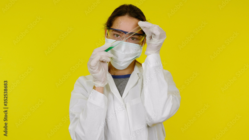 Nurse in protective medical mask, glasses, latex gloves and a test tube in her hands. Girl in a white coat on a yellow background