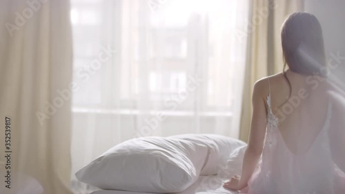  Rear view PAN of unrecognizable young woman in pink nightdress sitting on her bed on tranquil morning and stretching while enjoying sunlight