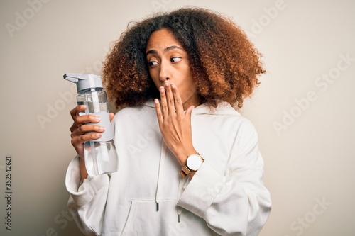 Young african american sportswoman with afro hair holding bottle of water cover mouth with hand shocked with shame for mistake, expression of fear, scared in silence, secret concept