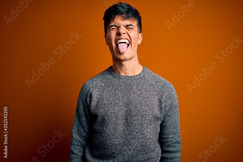 Young handsome man wearing casual sweater standing over isolated orange background sticking tongue out happy with funny expression. Emotion concept.