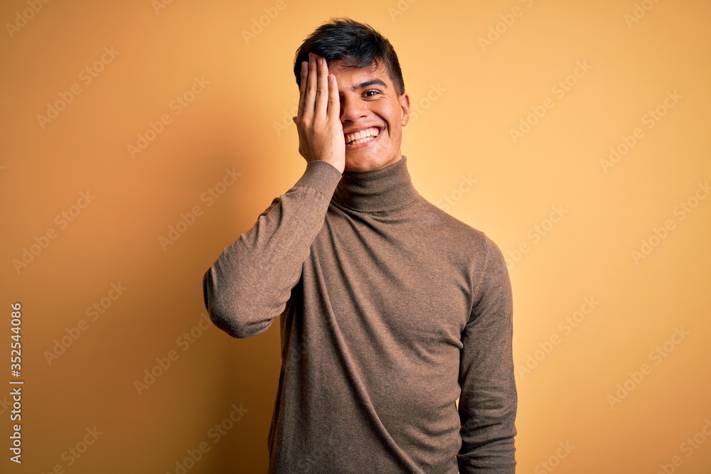 Young handsome man wearing casual turtleneck sweater over isolated yellow background covering one eye with hand, confident smile on face and surprise emotion. Young handsome man wearing casual turtleneck sweater over isolated yellow background covering one eye with hand, confident smile on face and surprise emotion.