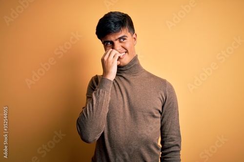 Young handsome man wearing casual turtleneck sweater over isolated yellow background looking stressed and nervous with hands on mouth biting nails. Anxiety problem.