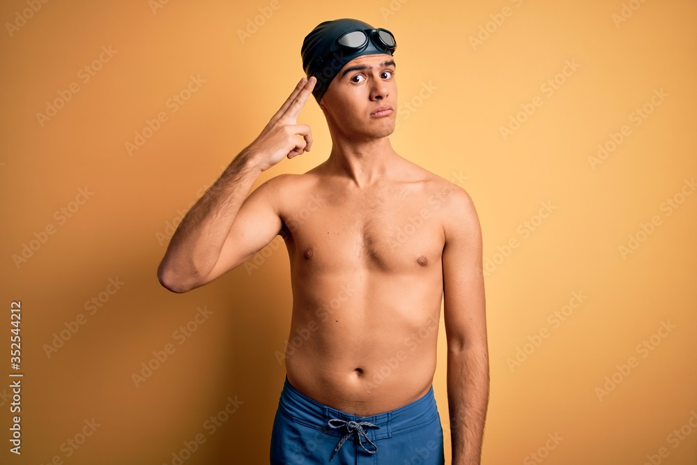 Young handsome man shirtless wearing swimsuit and swim cap over isolated yellow background Shooting and killing oneself pointing hand and fingers to head like gun, suicide gesture.
