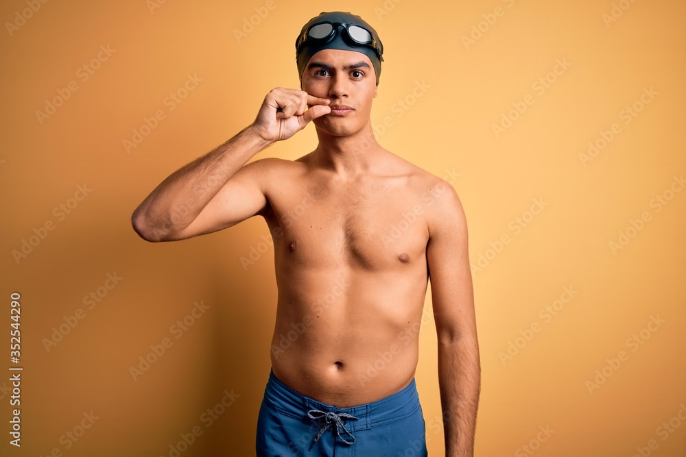 Young handsome man shirtless wearing swimsuit and swim cap over isolated yellow background mouth and lips shut as zip with fingers. Secret and silent, taboo talking