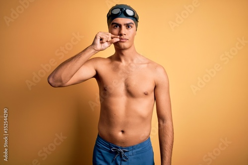 Young handsome man shirtless wearing swimsuit and swim cap over isolated yellow background mouth and lips shut as zip with fingers. Secret and silent, taboo talking © Krakenimages.com