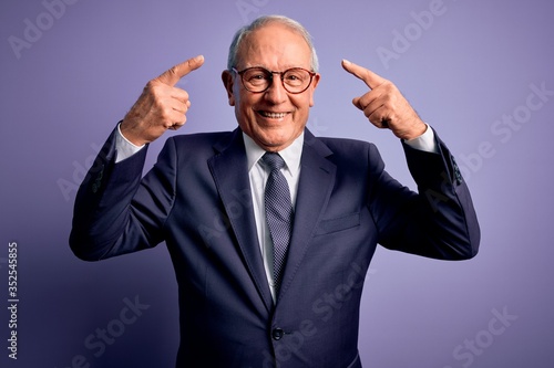 Grey haired senior business man wearing glasses and elegant suit and tie over purple background smiling pointing to head with both hands finger, great idea or thought, good memory