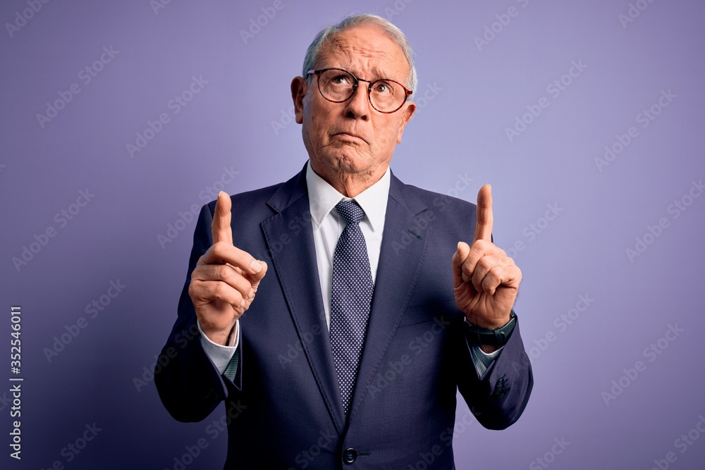 Grey haired senior business man wearing glasses and elegant suit and tie over purple background Pointing up looking sad and upset, indicating direction with fingers, unhappy and depressed. Grey haired senior business man wearing glasses and elegant suit and tie over purple background Pointing up looking sad and upset, indicating direction with fingers, unhappy and depressed.