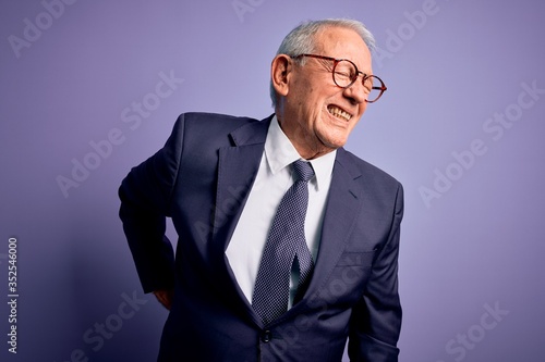 Grey haired senior business man wearing glasses and elegant suit and tie over purple background Suffering of backache, touching back with hand, muscular pain