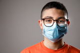 Ritratto di ragazzo giovane con occhiali da vista , maglia arancione e mascherina chirurgica , isolato su sfondo grigio