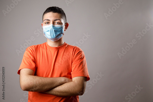 Ragazzo giovane con maglia arancione e mascherina chirurgica , isolato su sfondo grigio guarda con braccia incrociate photo