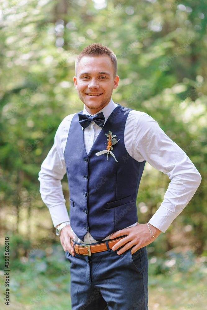 A serious portrait of a handsome groom in a blue suit and bow tie is standing against the backdrop of greenery in the forest. A serious portrait of a handsome groom in a blue suit and bow tie is standing against the backdrop of greenery in the forest.