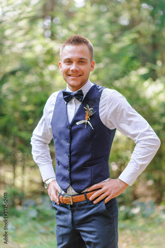 A serious portrait of a handsome groom in a blue suit and bow tie is standing against the backdrop of greenery in the forest.