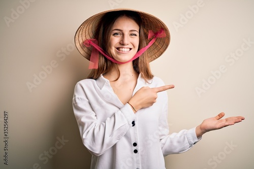 Young beautiful redhead woman wearing asian traditional conical hat over white background amazed and smiling to the camera while presenting with hand and pointing with finger.