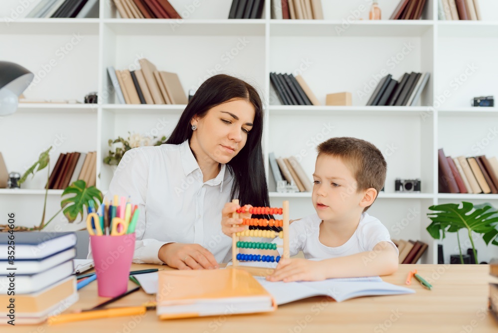 Female private tutor helping young student with homework at desk in bright child's room