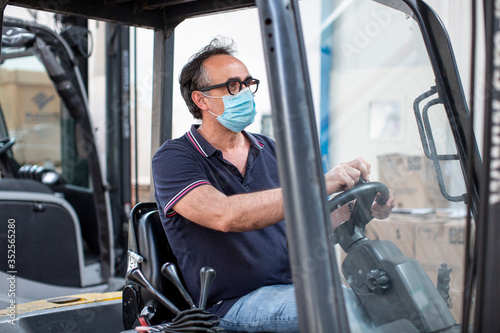 Operaio  con mascherina di protezione guida un carrello elevatore nel magazzino in cui lavora photo