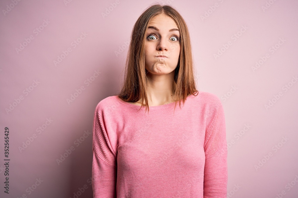 Obraz premium Young beautiful redhead woman wearing casual sweater over isolated pink background puffing cheeks with funny face. Mouth inflated with air, crazy expression.
