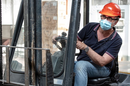 Operaio  con caschetto protettivo e mascherina di protezione guida un carrello elevatore nel magazzino in cui lavora photo