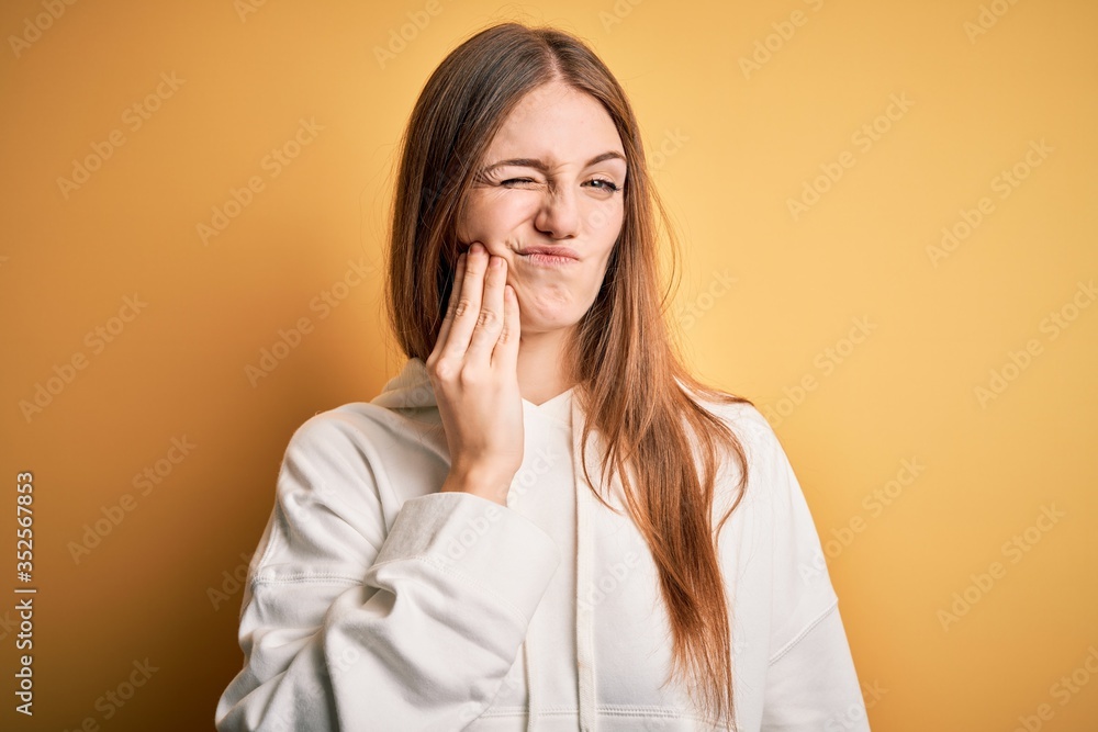 Young beautiful redhead sporty woman wearing sweatshirt over isolated yellow background touching mouth with hand with painful expression because of toothache or dental illness on teeth. Dentist Young beautiful redhead sporty woman wearing sweatshirt over isolated yellow background touching mouth with hand with painful expression because of toothache or dental illness on teeth. Dentist