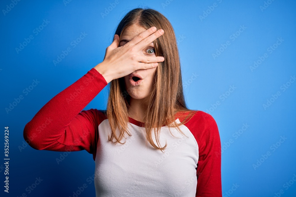 Young beautiful redhead woman wearing casual t-shirt over isolated blue background peeking in shock covering face and eyes with hand, looking through fingers with embarrassed expression. Young beautiful redhead woman wearing casual t-shirt over isolated blue background peeking in shock covering face and eyes with hand, looking through fingers with embarrassed expression.