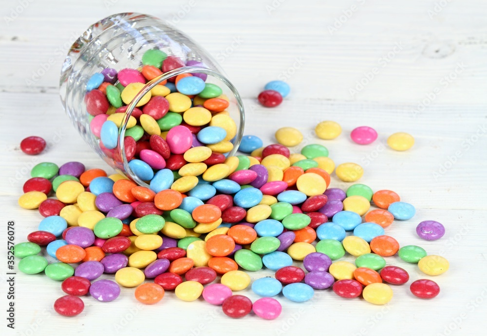 Glass full of colorful and sweet smarties. Chocolate buttons in colors of rainbow. Delicious candies on white wooden table. Glass full of colorful and sweet smarties. Chocolate buttons in colors of rainbow. Delicious candies on white wooden table.