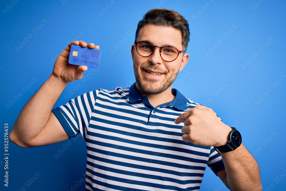 Young man with blue eyes wearing glasses and holding credit card over blue background with surprise face pointing finger to himself Young man with blue eyes wearing glasses and holding credit card over blue background with surprise face pointing finger to himself