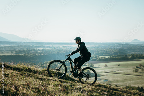 Fototapeta Naklejka Na Ścianę i Meble -  Young athletic man on an e-bike pedaling up the hill with a beautiful view. Amazing nature for meditating, hiking, biking and exploring.
