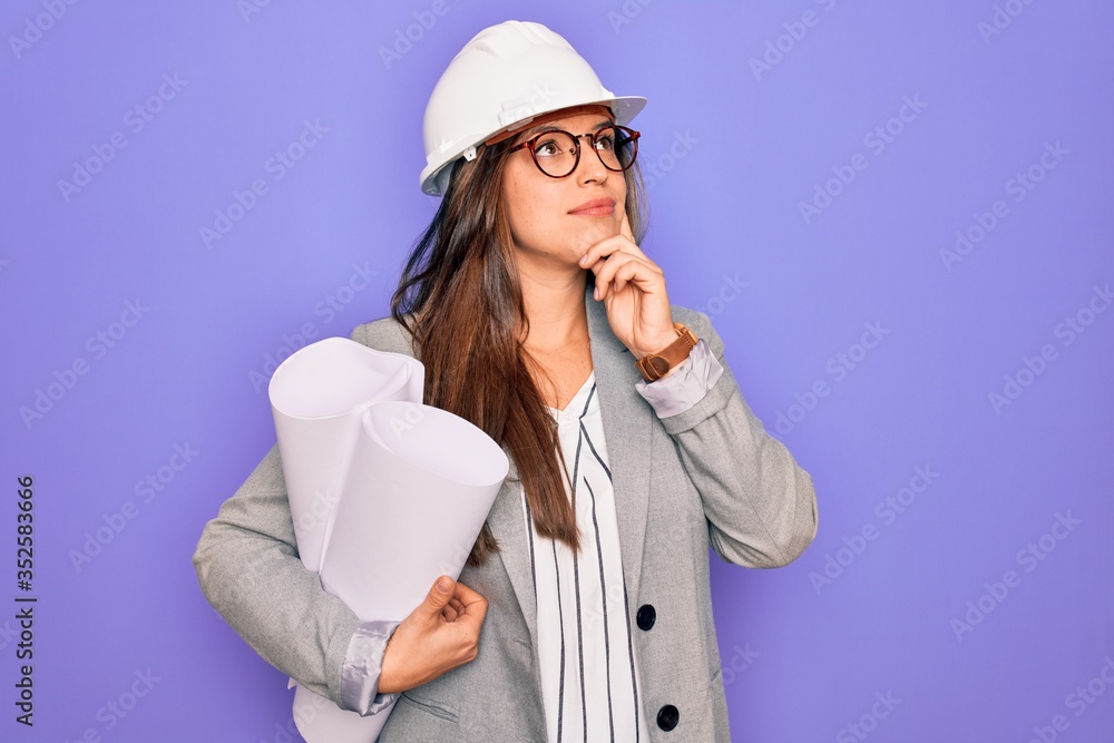 Young hispanic architect woman wearing builder hat and holding blueprints over yellow background serious face thinking about question, very confused idea Young hispanic architect woman wearing builder hat and holding blueprints over yellow background serious face thinking about question, very confused idea