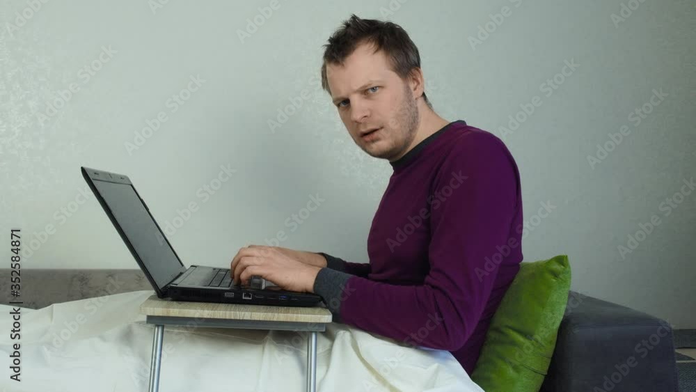 Young unshaven man working on a laptop while lying in bed and looking at the camera, internet. Work at home, freelance, technology Young unshaven man working on a laptop while lying in bed and looking at the camera, internet. Work at home, freelance, technology