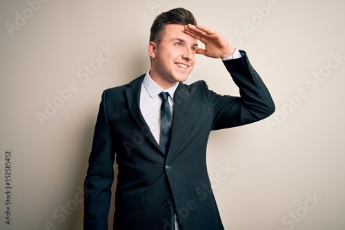 Young handsome business man wearing elegant suit and tie over isolated background very happy and smiling looking far away with hand over head. Searching concept.