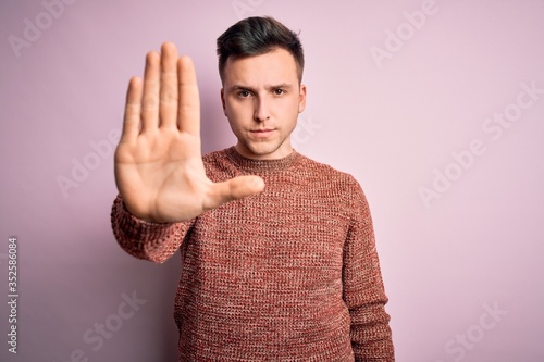 Young handsome caucasian man wearing casual winter sweater over pink isolated background doing stop sing with palm of the hand. Warning expression with negative and serious gesture on the face.