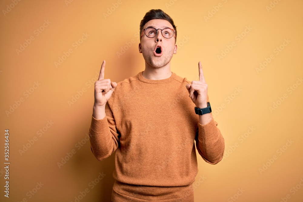 Young handsome caucasian man wearing glasses and casual winter sweater over yellow background amazed and surprised looking up and pointing with fingers and raised arms. Young handsome caucasian man wearing glasses and casual winter sweater over yellow background amazed and surprised looking up and pointing with fingers and raised arms.